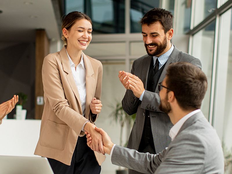 A woman in a business suit shakes hands with a man while two others smile and applaud in a modern office setting.