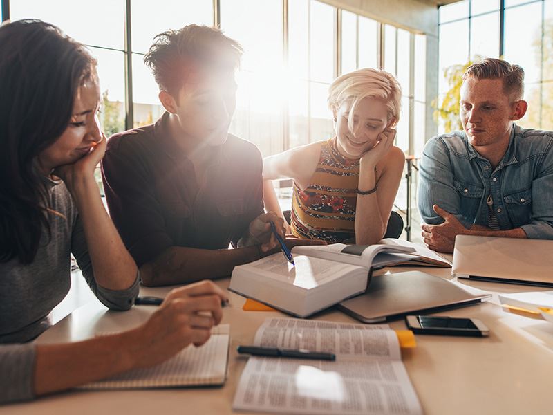Four young people are studying together at a table, engaging with a book.